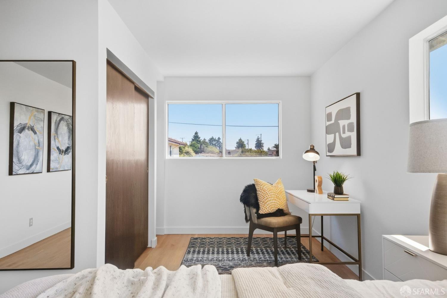 Staged bedroom with a desk and chair beside a large sunny window.