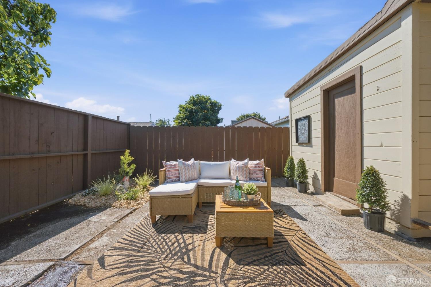 Fenced backyard patio with wicker seating, a patterned rug, and a light-colored outbuilding.