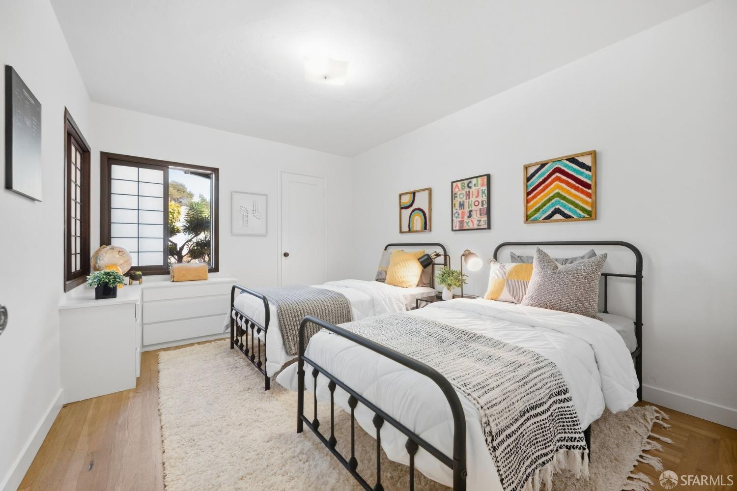 Bedroom with two black metal twin beds, colorful pillows, framed artwork, and a window overlooking greenery.