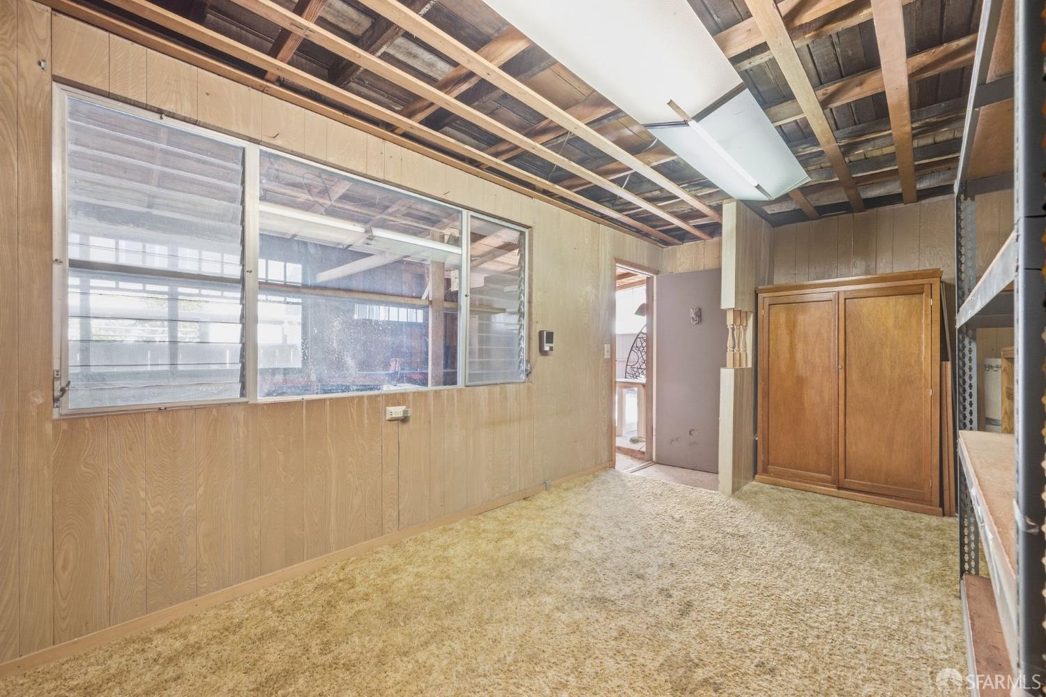 Empty carpeted room with wood-paneled walls, exposed ceiling rafters, and a large interior window.