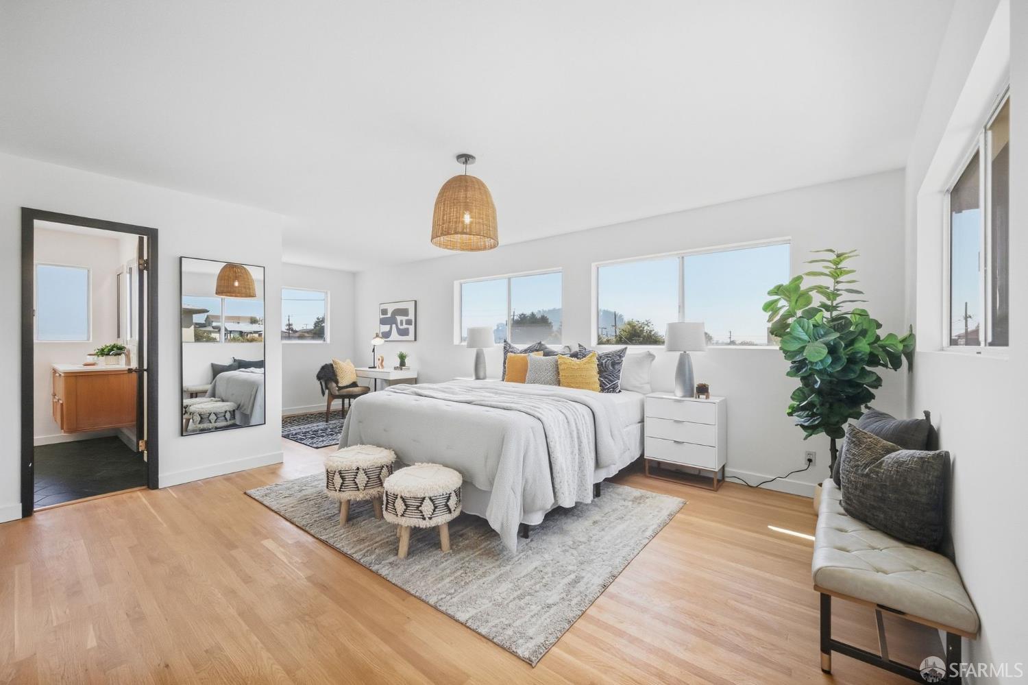 Staged bedroom with a white bed, woven pendant light, light wood floors, and windows along the back wall.