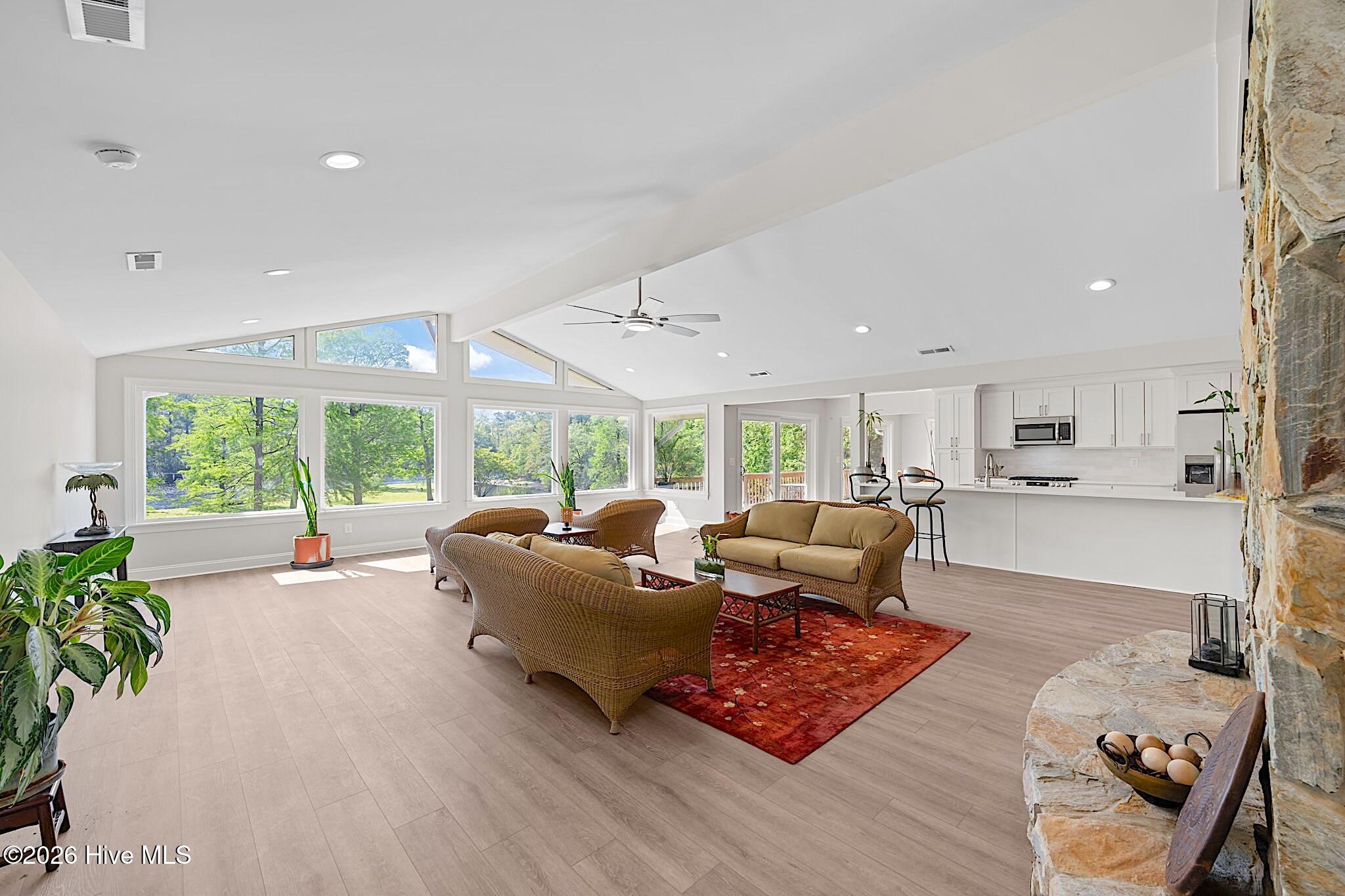 Furnished living room with expansive windows, wicker seating, vaulted ceiling, and stone fireplace beside an open kitchen.