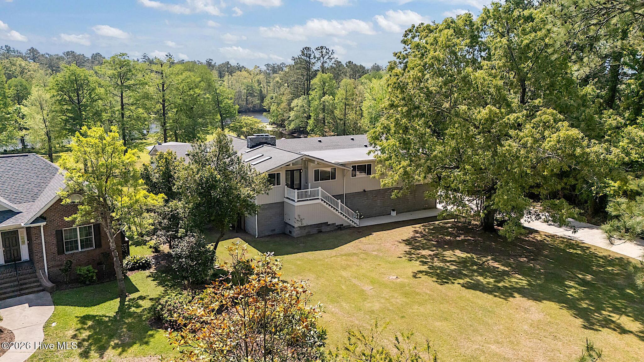 Raised home with exterior stairs and broad lawn, surrounded by mature trees with water visible beyond.