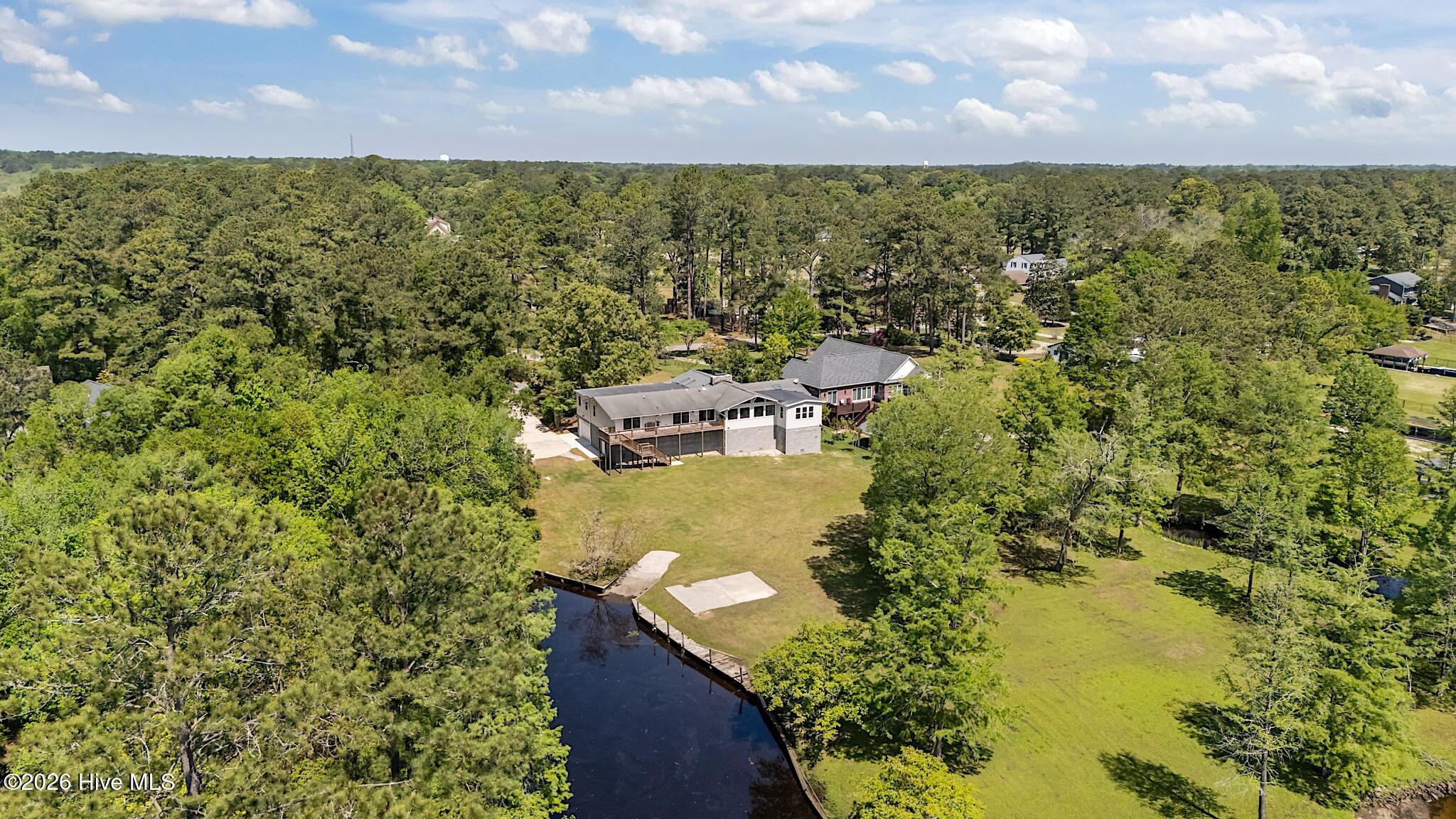 Aerial view of a home beside a narrow waterway, with a broad lawn and wooded surroundings.