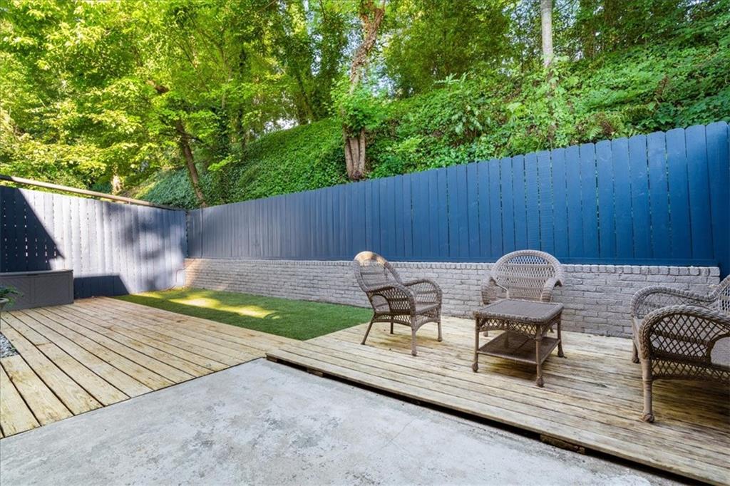 Fenced backyard patio with a wood deck, wicker seating, artificial turf, and leafy trees.