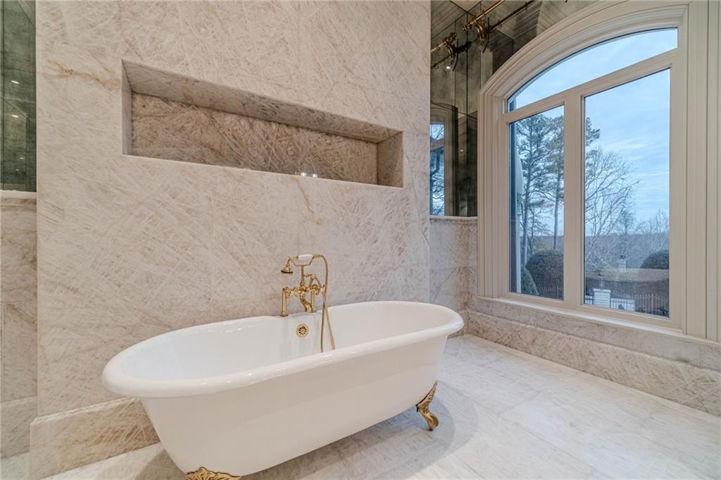 Marble bathroom with a freestanding clawfoot tub, gold fixtures, and a large arched window.