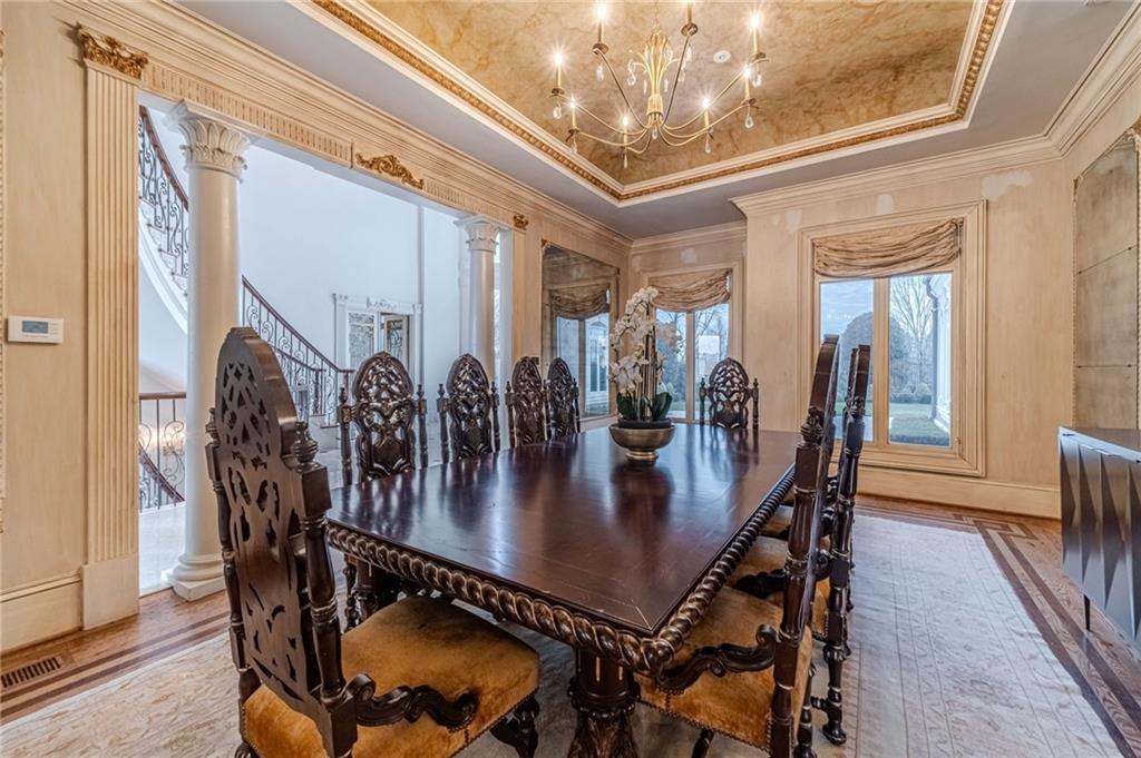 Formal dining room with an ornate carved table, chandelier, and columns beside an open staircase.