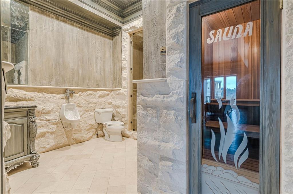 Stone bathroom with a glass-front sauna, toilet, urinal, and ornate vanity.