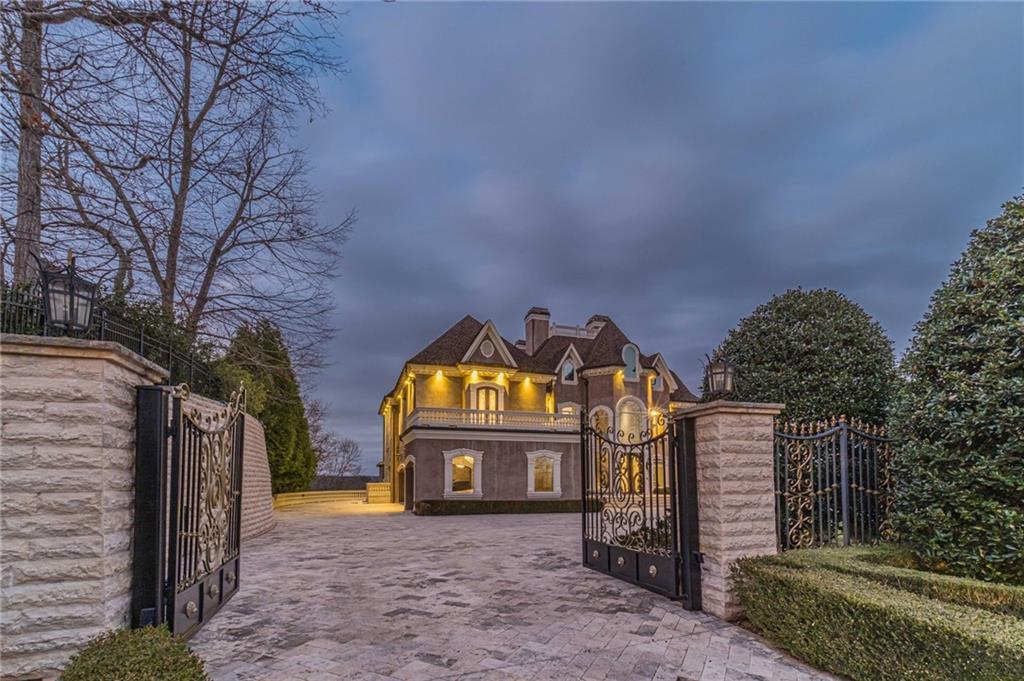Brick residence behind an ornate open gate, with a stone-paved driveway and glowing exterior lights.
