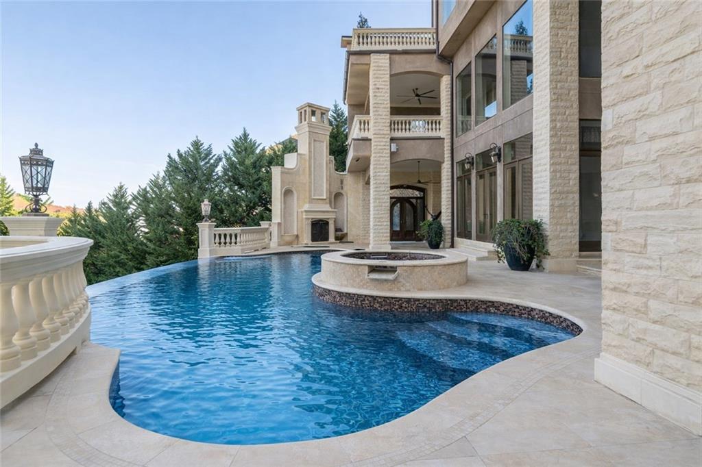 Curved swimming pool and raised spa beside a stone house with balconies and an outdoor fireplace.
