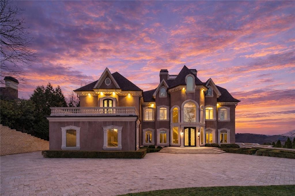 Large stone mansion with illuminated windows and a broad paved driveway beneath a pink-purple dusk sky.