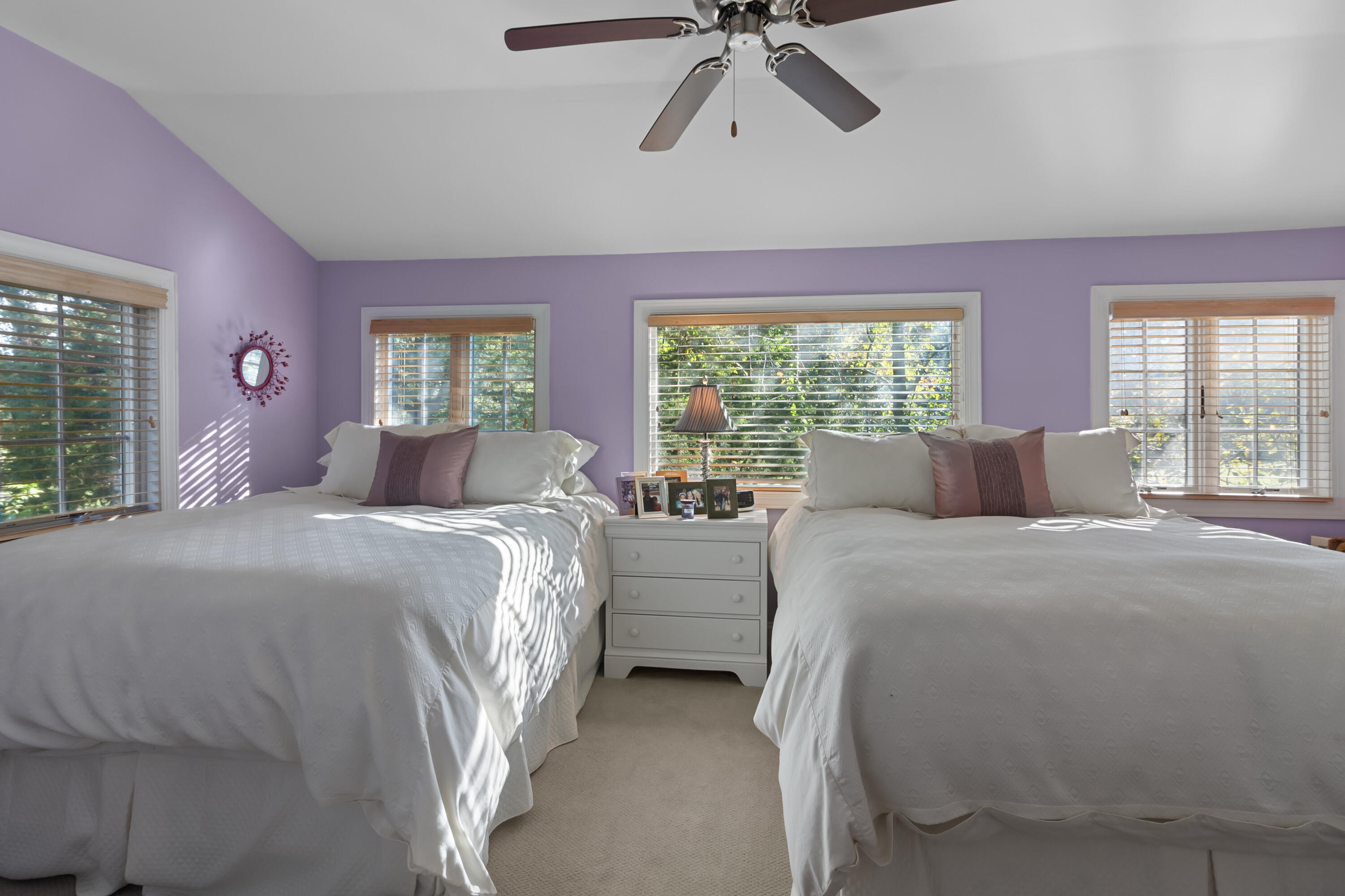 Bedroom with two white beds, purple walls, and windows overlooking trees.
