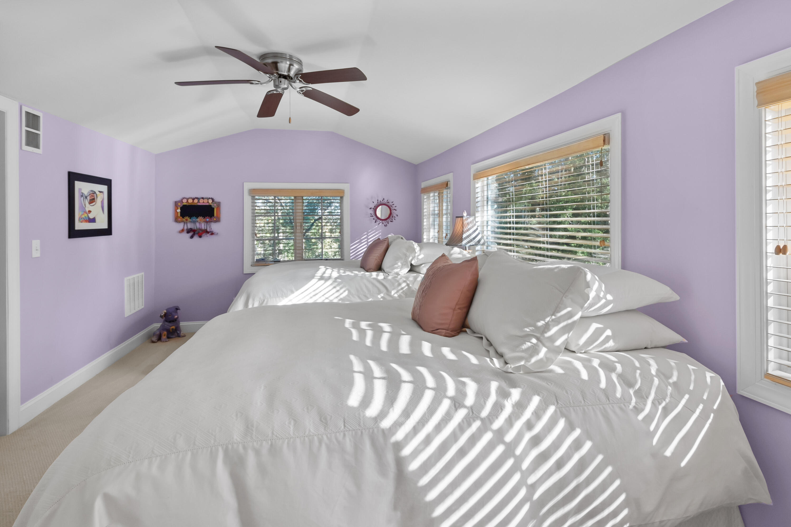 Bedroom with lavender walls, vaulted ceiling, and sunlight casting striped shadows across a large white bed.