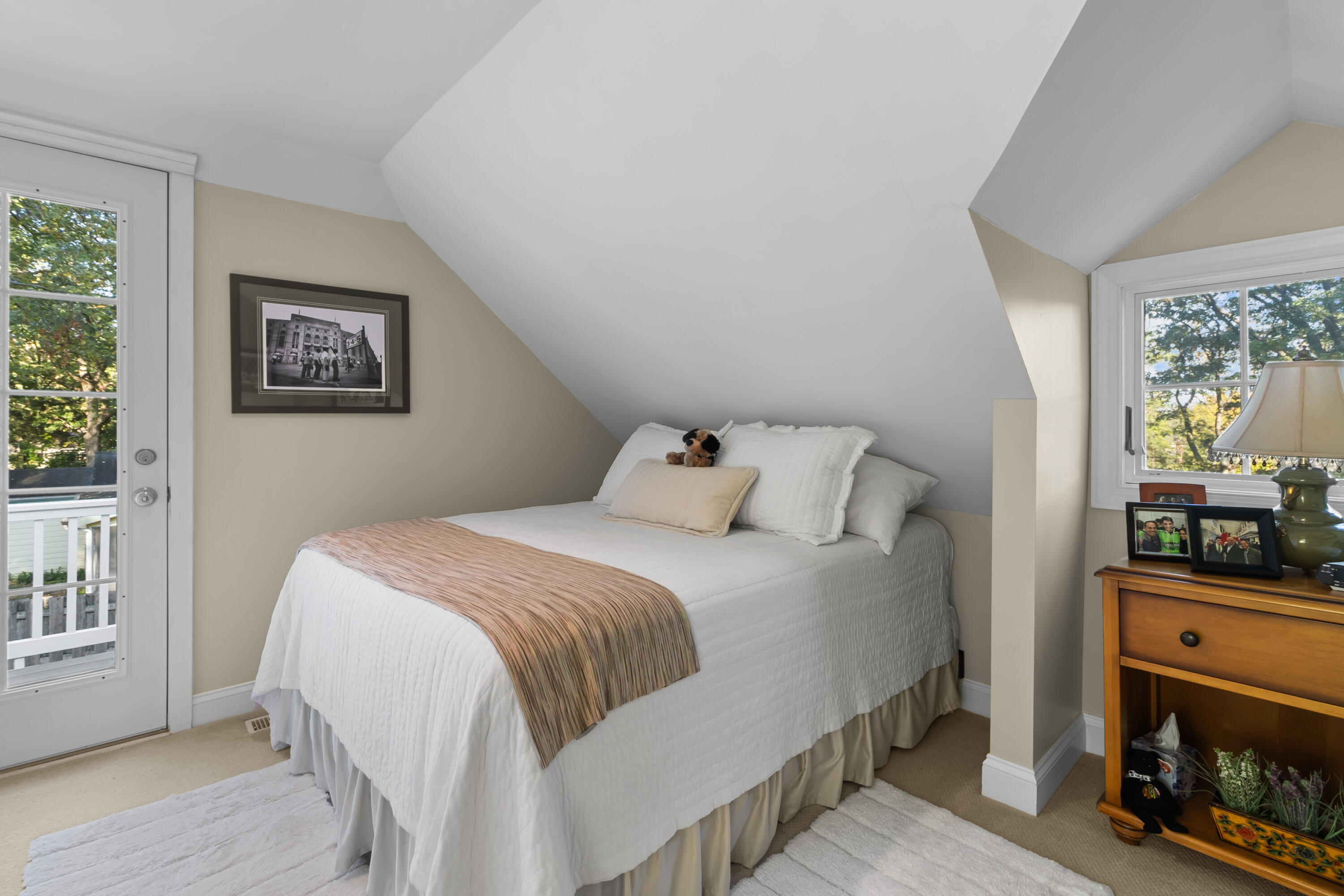 Attic bedroom with a white bed beneath a sloped ceiling, beside a glass door and leafy windows.