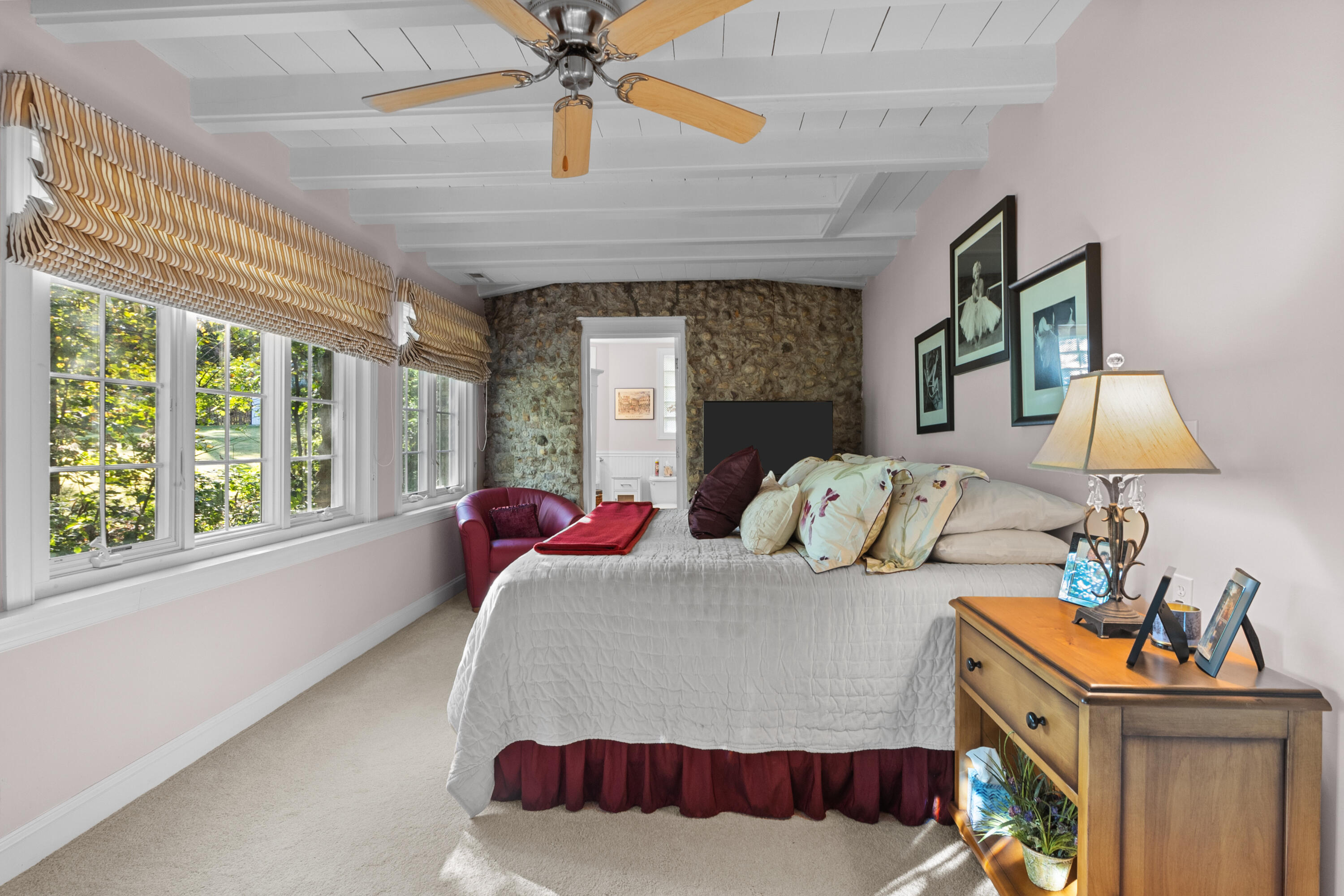 Bedroom with a stone accent wall, white beamed ceiling, and sunlit windows with striped shades.