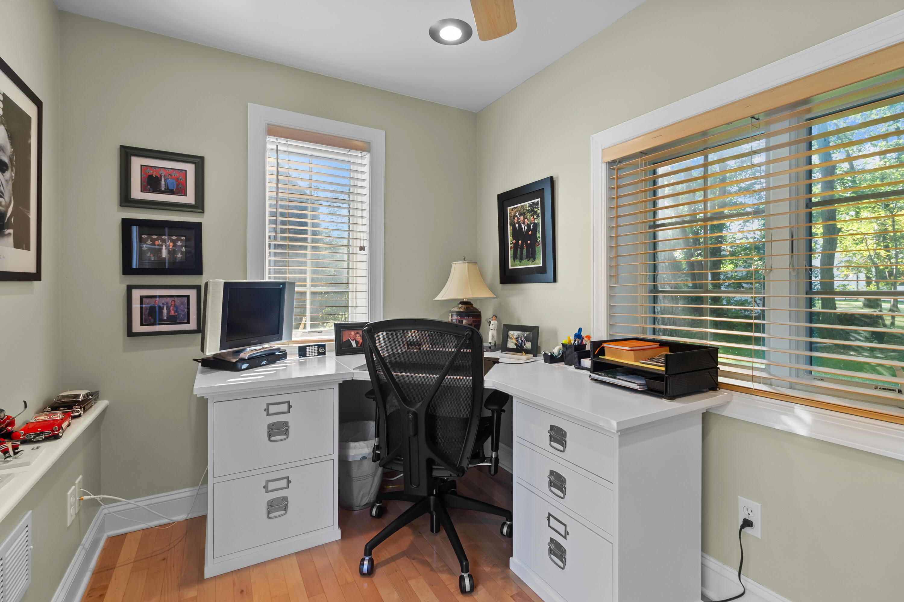 Home office with a white corner desk, black mesh chair, and large windows overlooking trees.