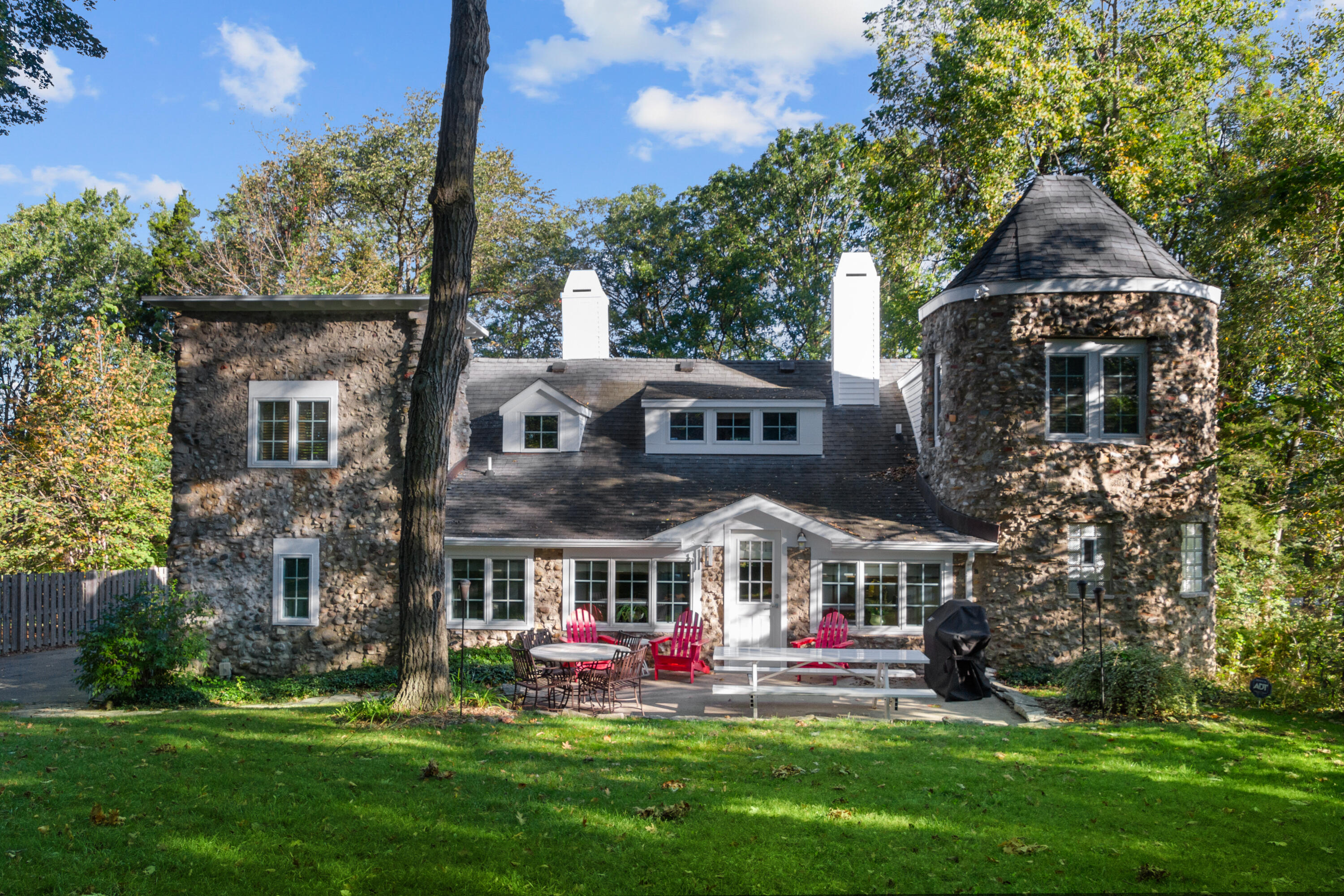Stone house rear exterior with round tower and patio seating beneath mature trees.