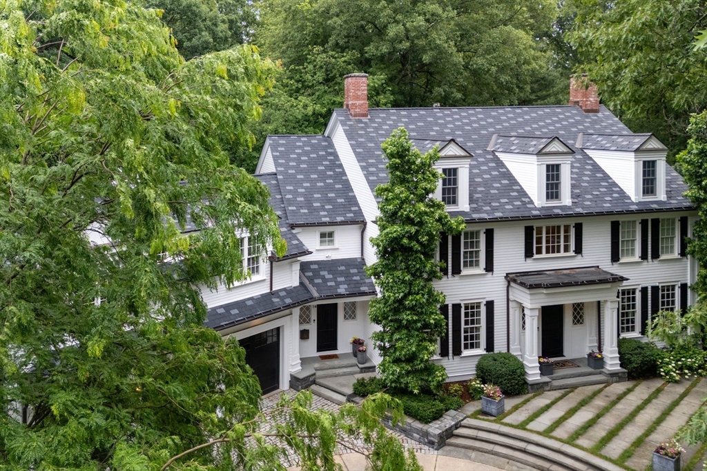 White clapboard house with black shutters, dormers, brick chimneys, and a landscaped stone driveway.