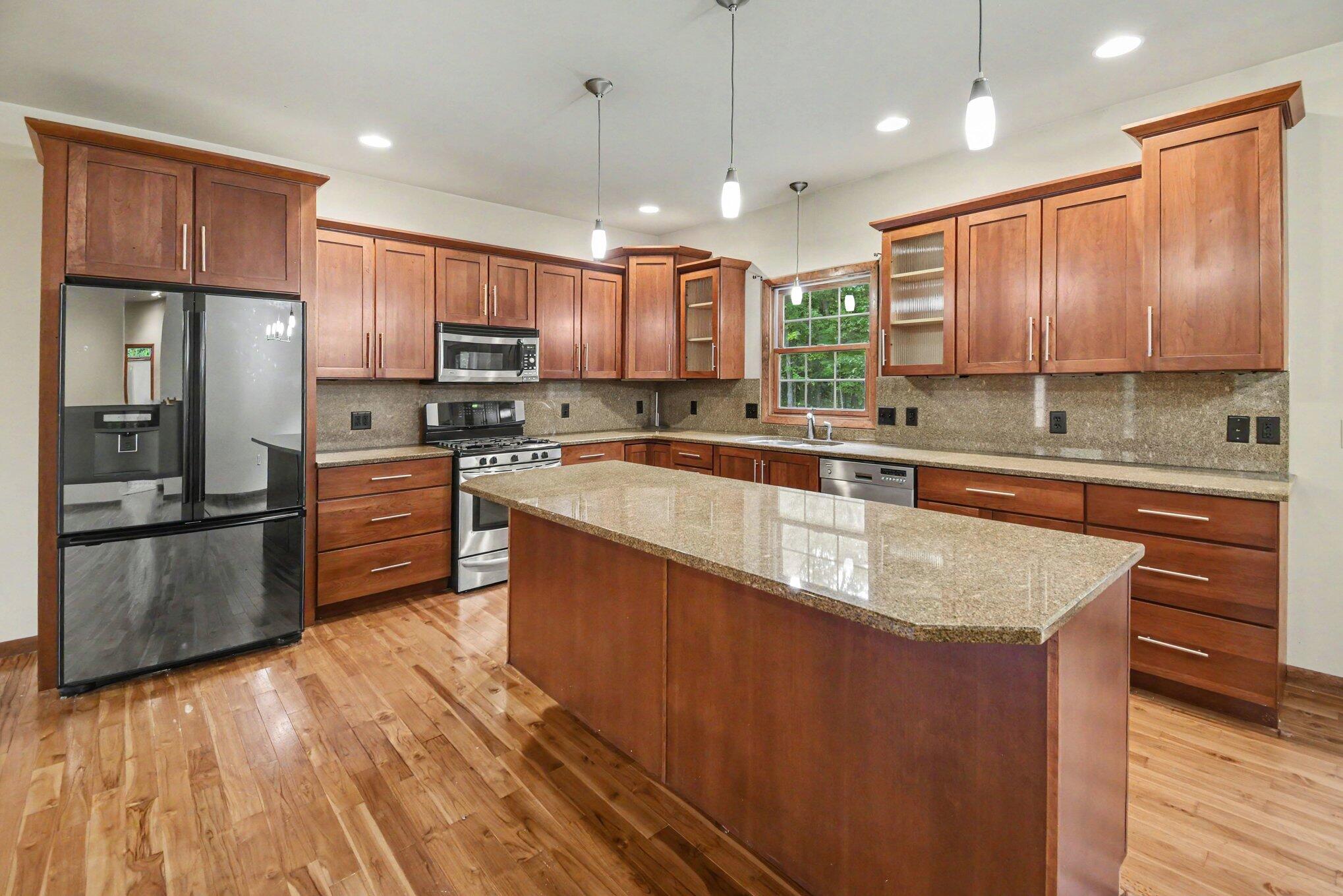 Kitchen with a large granite island, cherry-toned cabinets, stainless steel appliances, and hardwood floors.