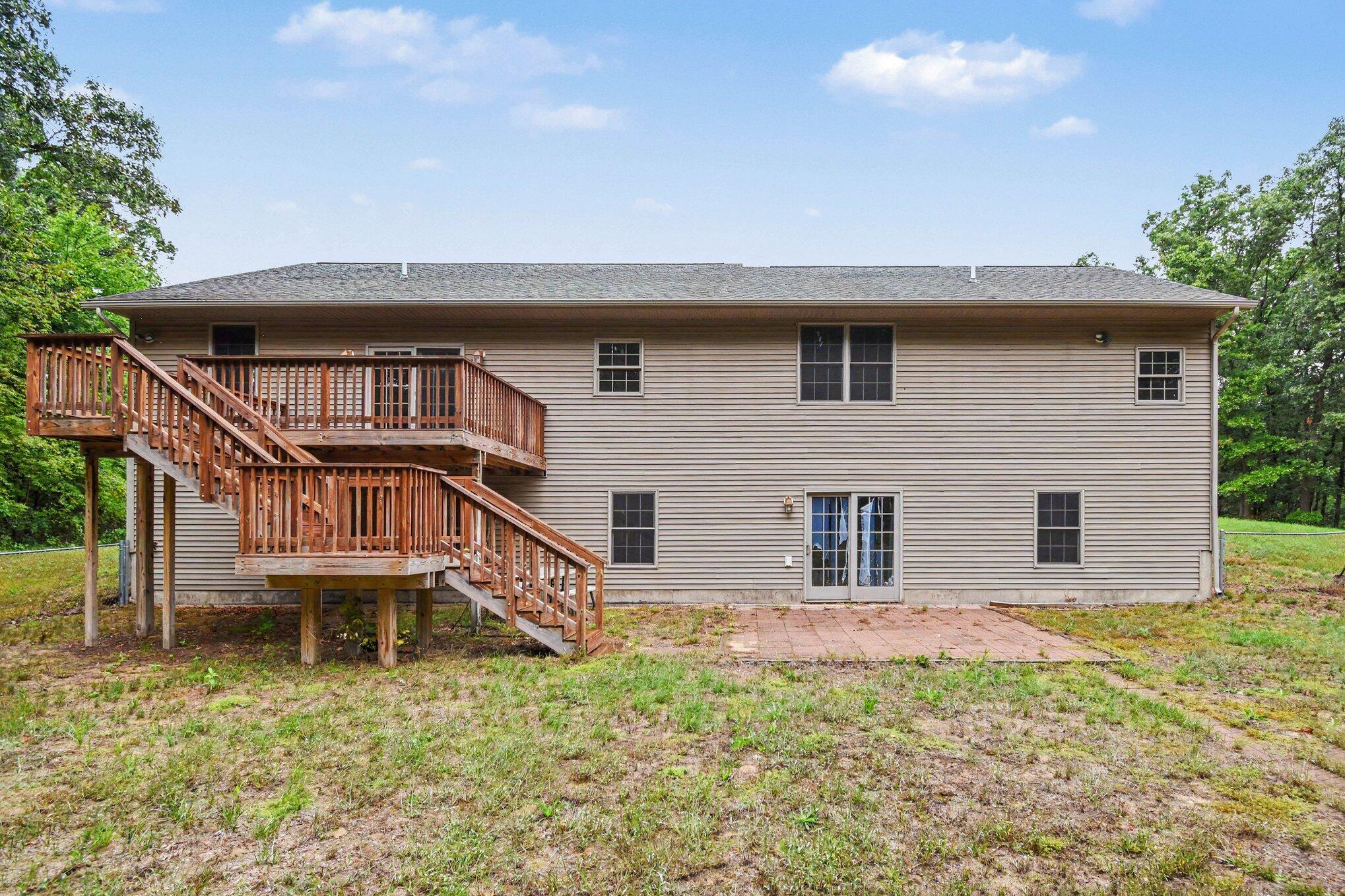 Rear of a beige-sided home with a raised wooden deck, long staircase, and brick patio.