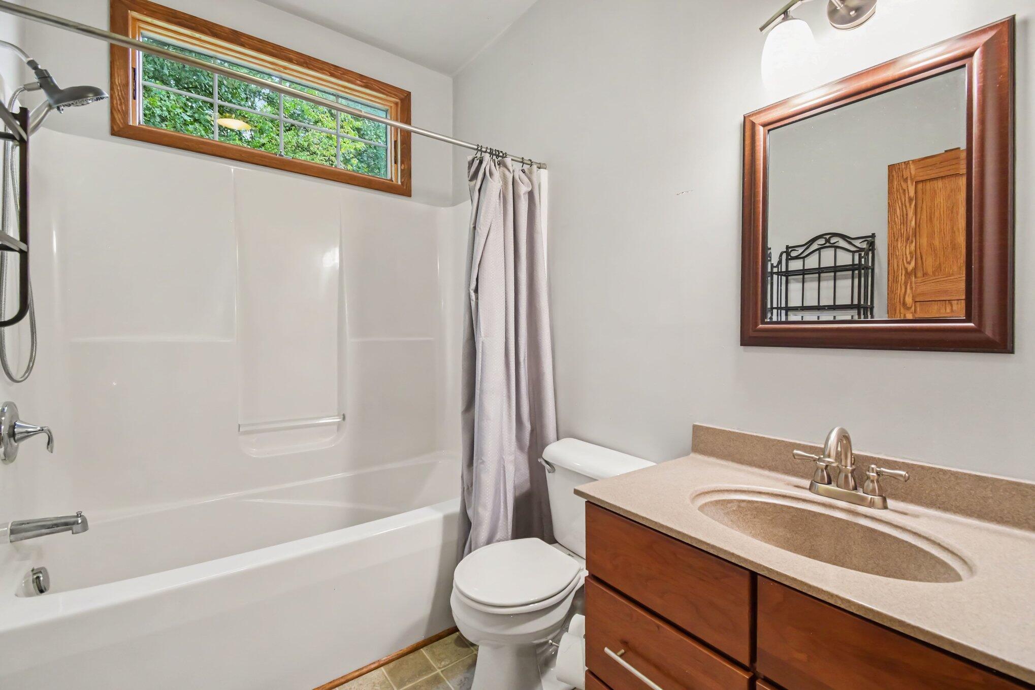 Bathroom with a white tub and shower, wood-trimmed window, and wood vanity with a beige countertop.