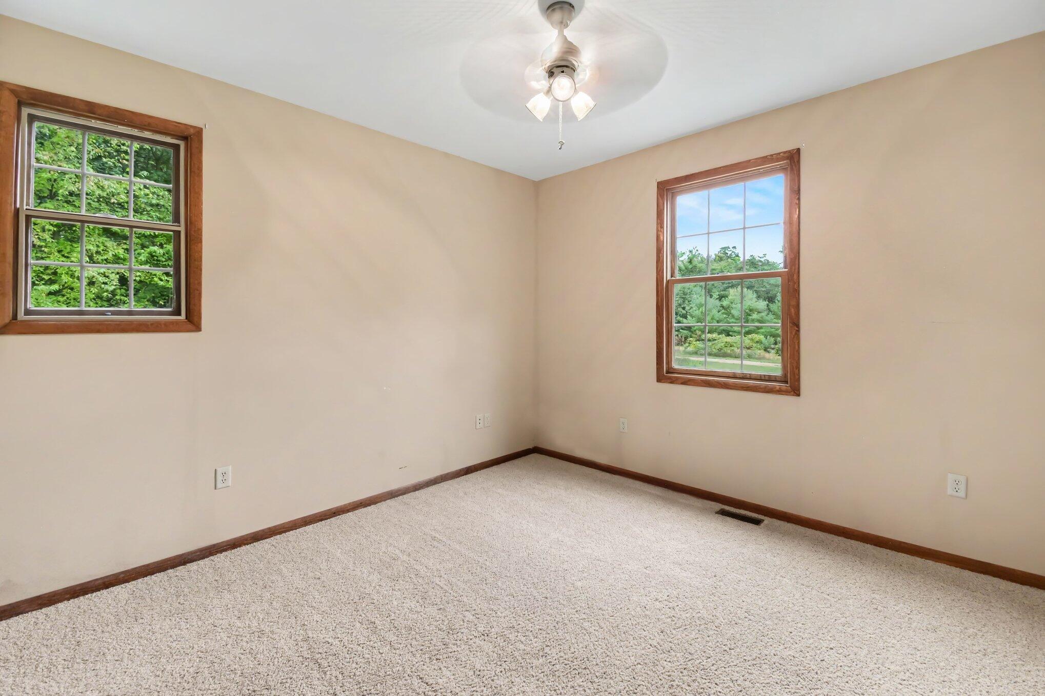 Empty bedroom with beige walls, carpet, two wood-trimmed windows, and a ceiling fan.