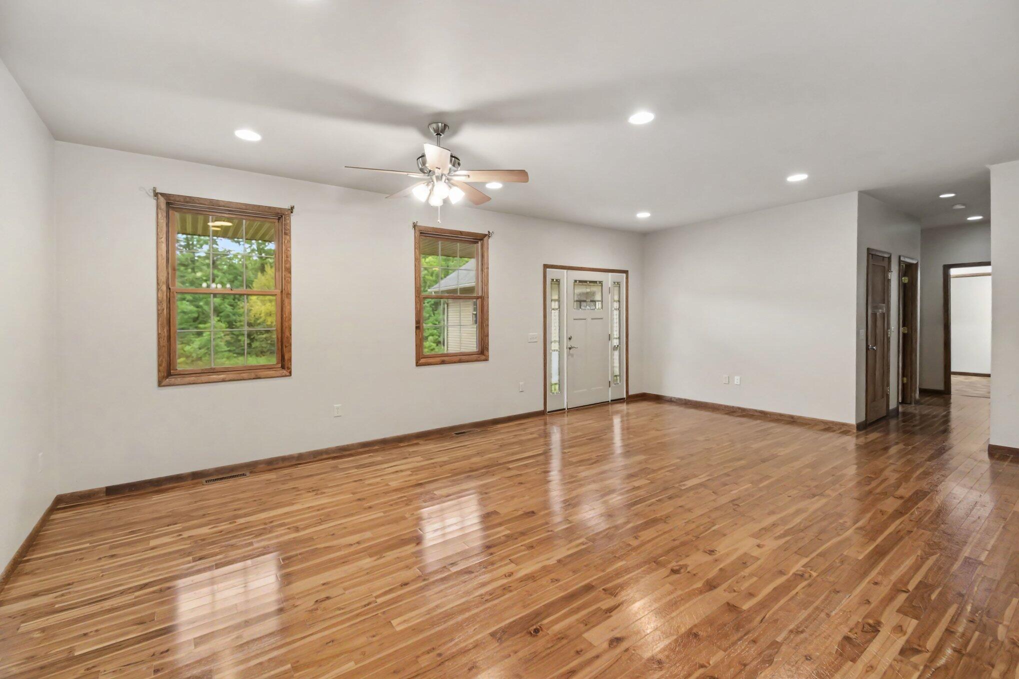 Empty living room with glossy hardwood floors, wood-trimmed windows, and a ceiling fan.