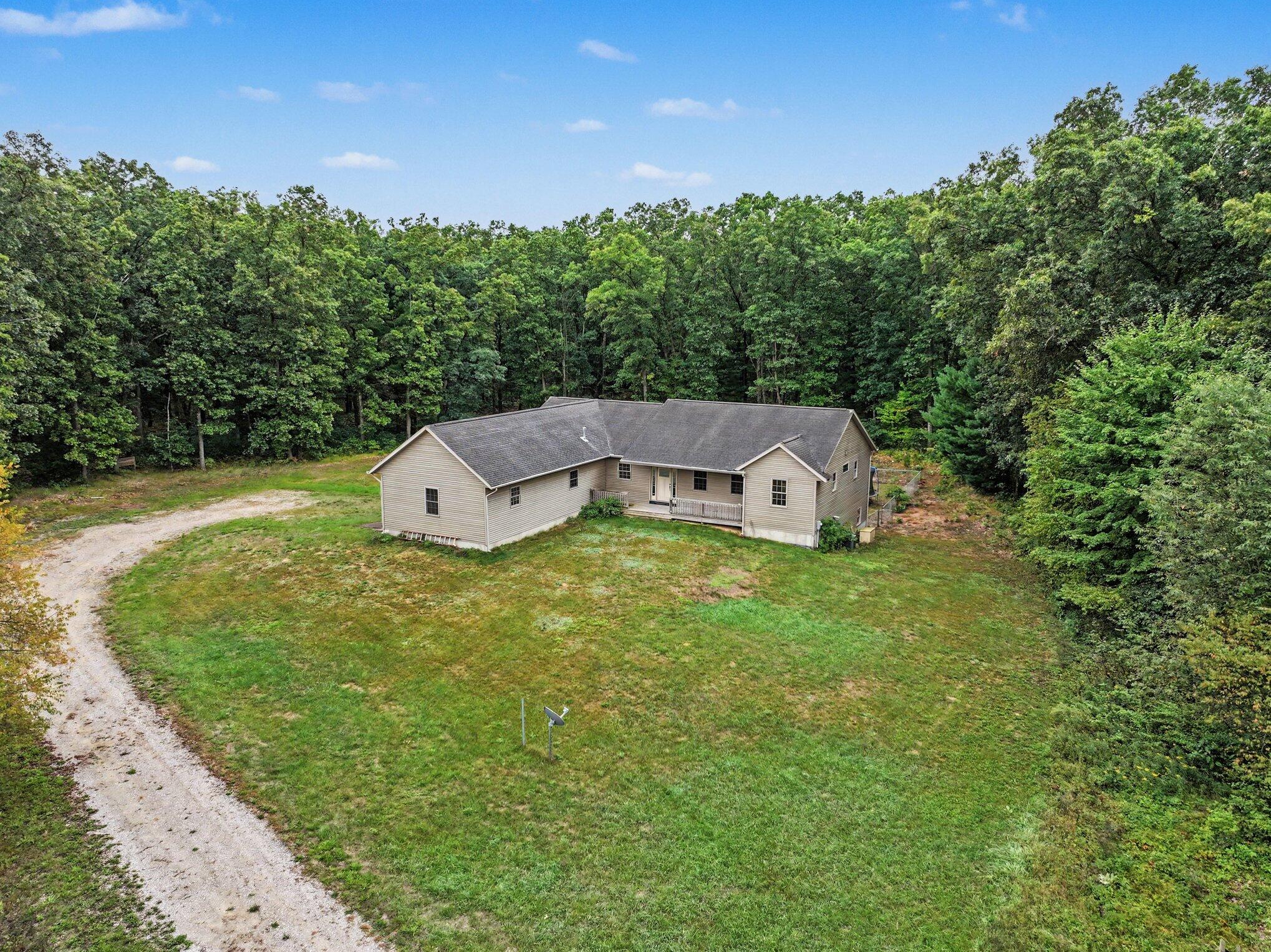 Aerial view of a beige home nestled among trees, with a gravel driveway and spacious lawn.