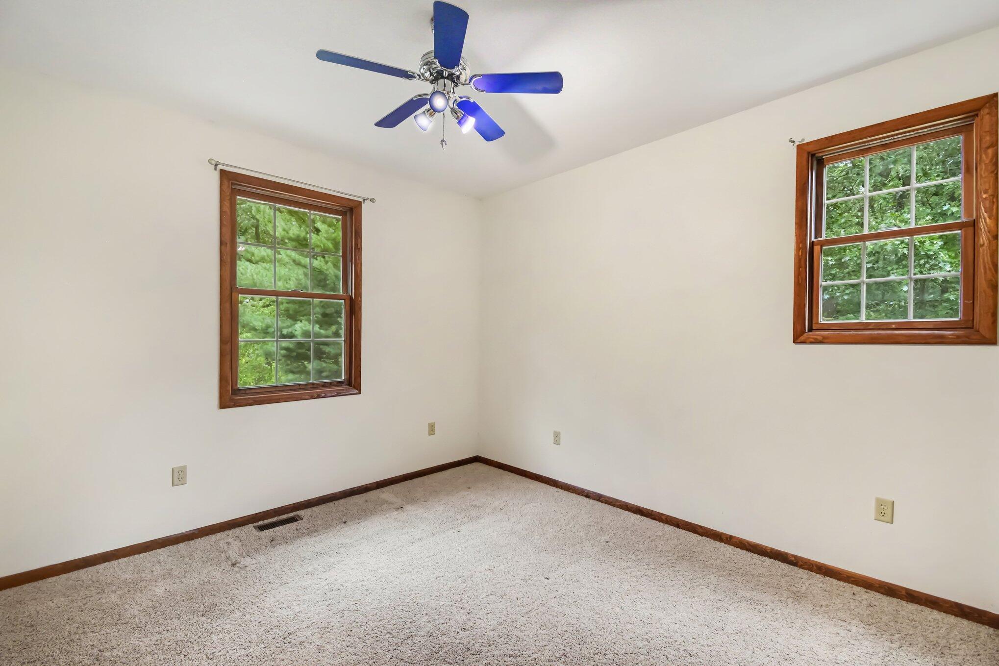 Empty carpeted room with two wood-trimmed windows and a blue ceiling fan.