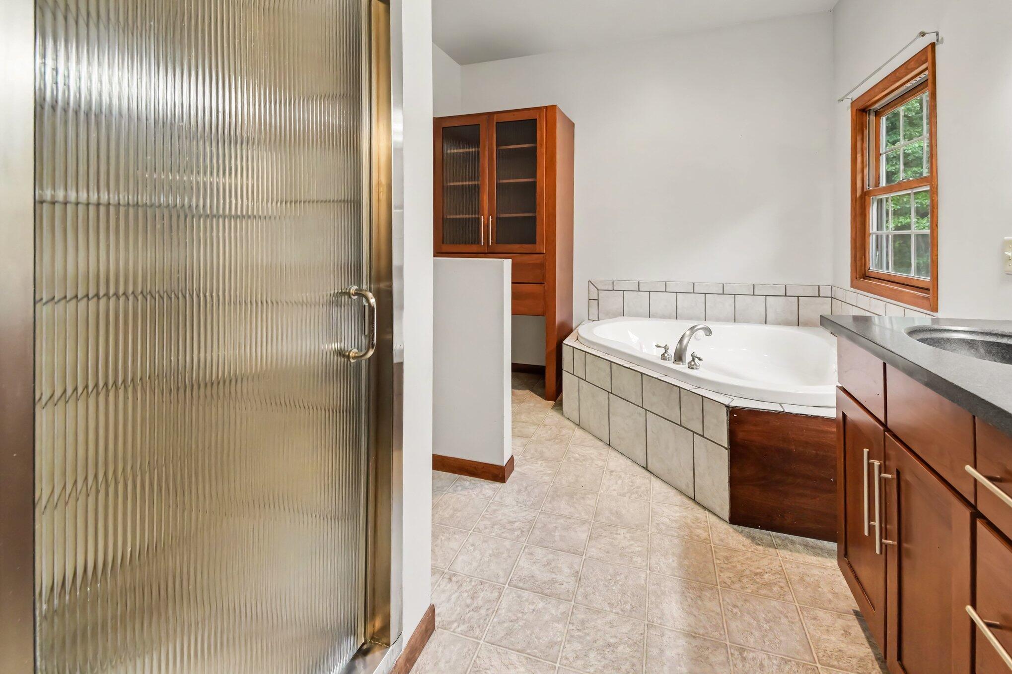 Bathroom with a tiled corner soaking tub, wood cabinetry, and a ribbed glass shower door.