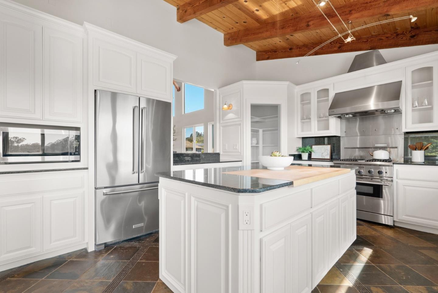 White kitchen with a large island, stainless steel appliances, slate floor, and exposed wood ceiling beams.