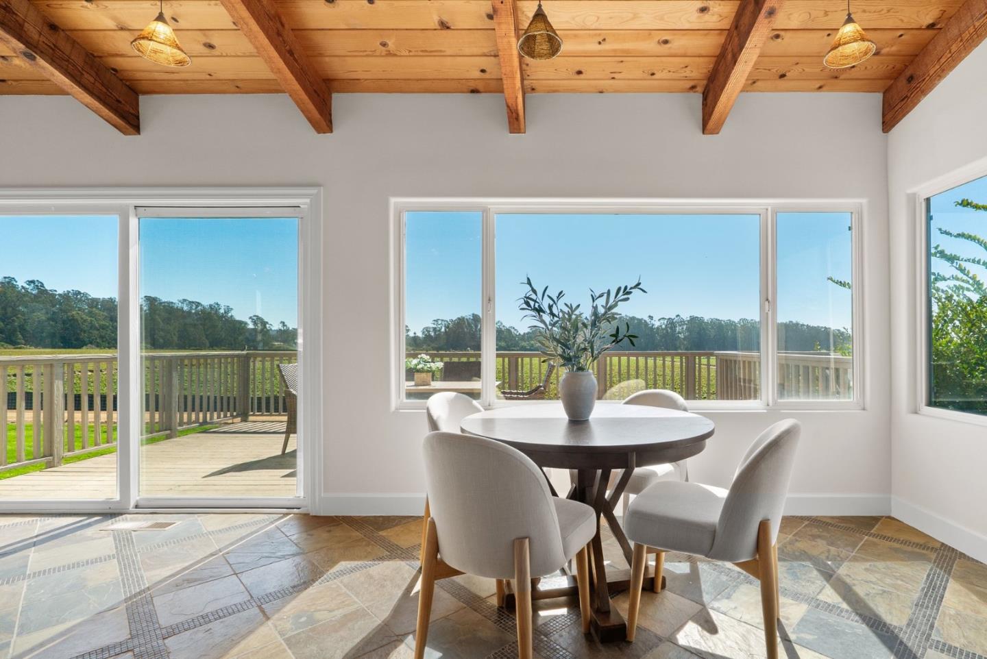 Dining area with a round table, exposed wood ceiling, and windows overlooking a deck and green fields.