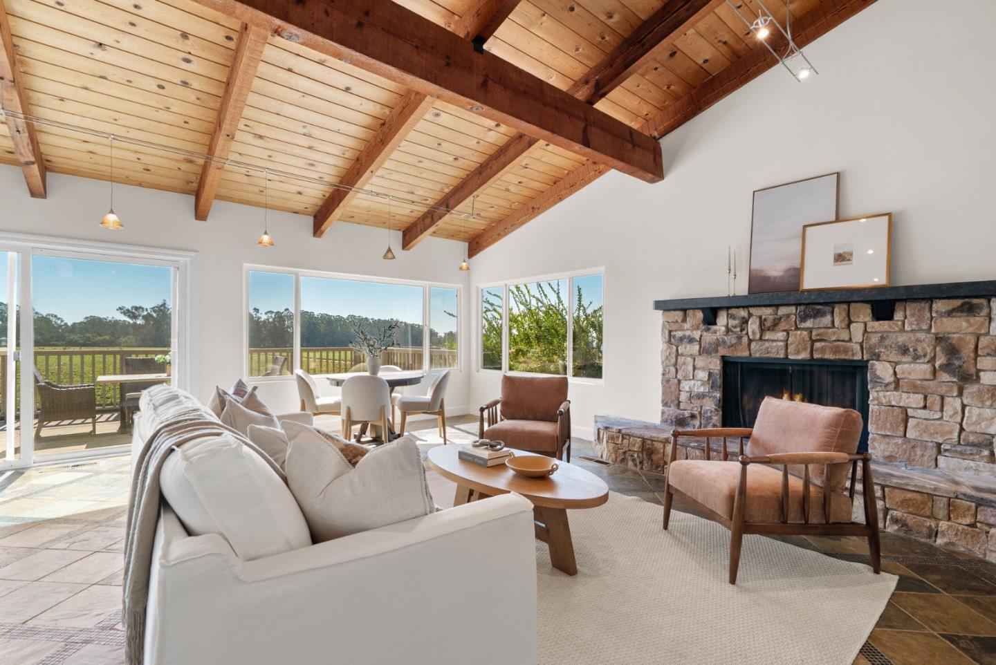 Living room with a vaulted wood ceiling, stone fireplace, and windows overlooking a deck and green landscape.
