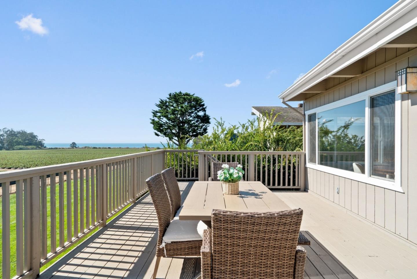 Furnished deck overlooking a field and ocean, with wicker dining chairs and a wooden table.