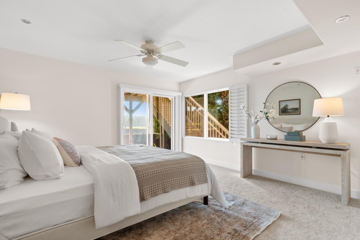 Bright bedroom with a white bed, round wall mirror, and sliding glass doors beside a wooden staircase.