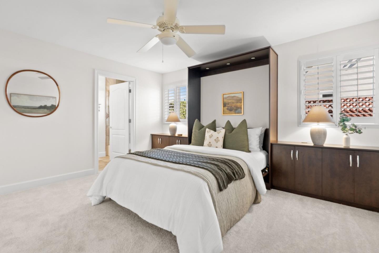 Staged bedroom with a white bed, dark wood built-ins, plantation shutters, and a ceiling fan.