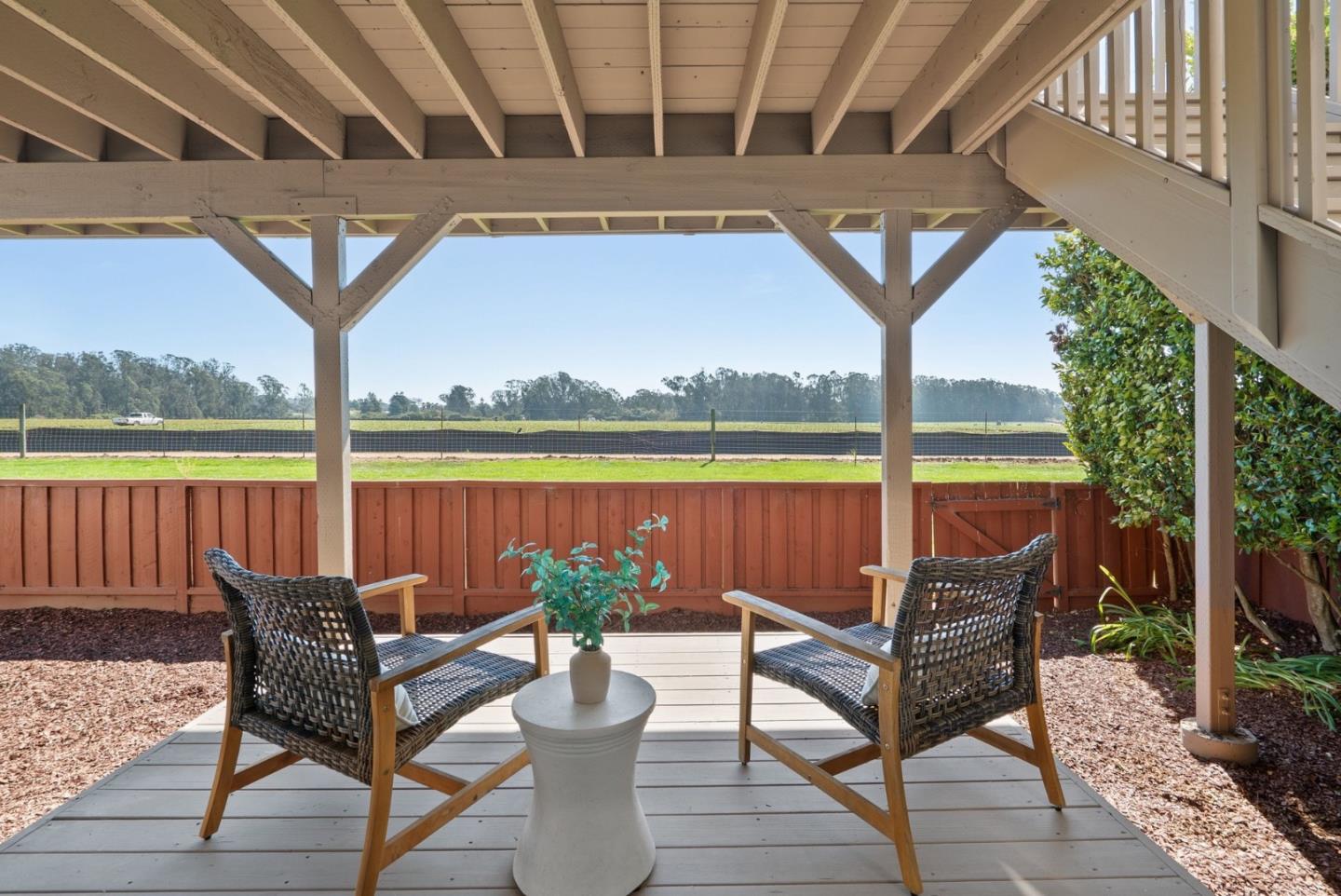Covered patio with two woven chairs, a small table, and open fields beyond a red fence.
