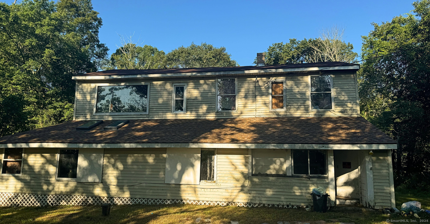 Home exterior with light siding, a broad lower roof, and mature trees surrounding it.