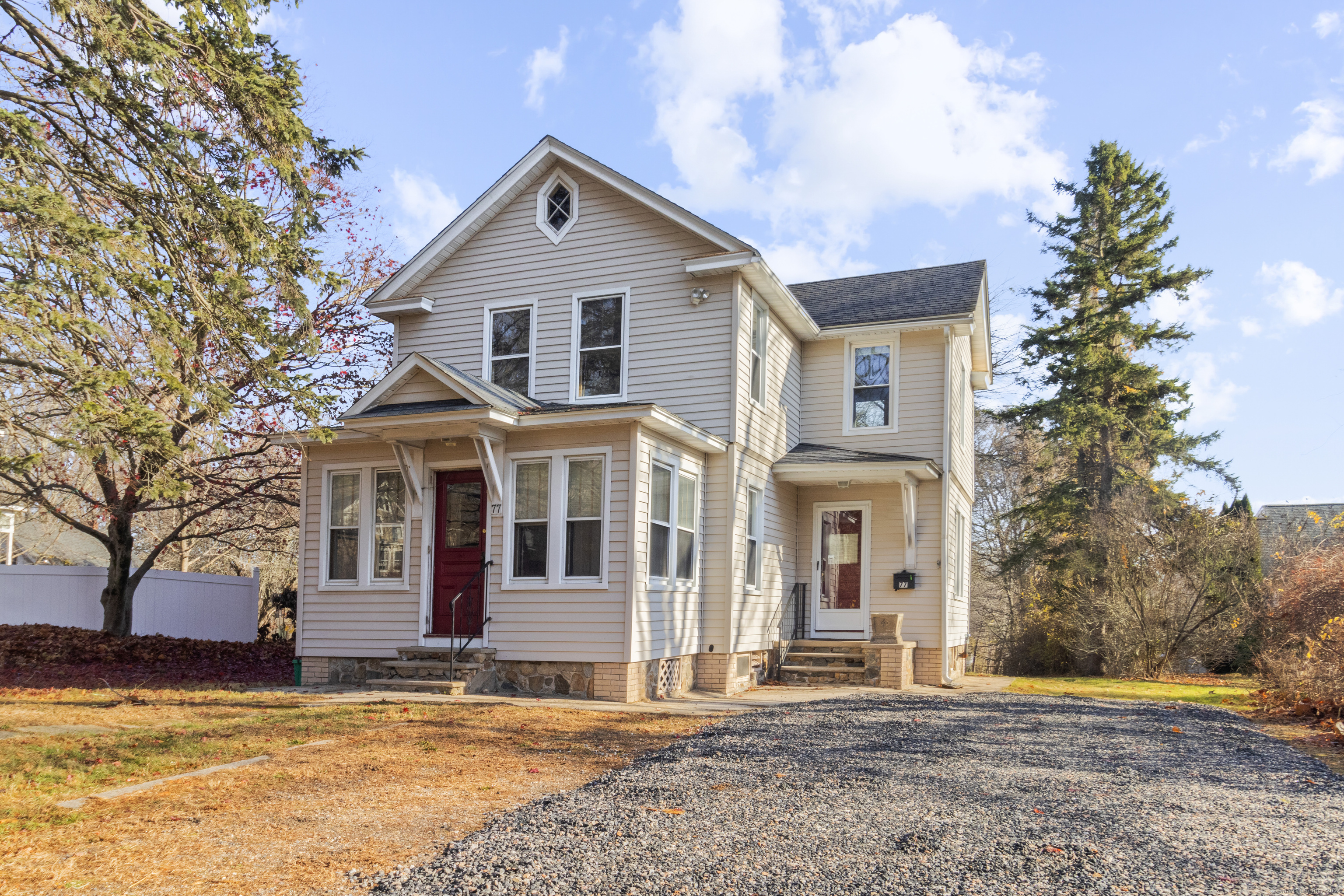 Two-story light-sided house with two red entry doors and a broad gravel driveway.