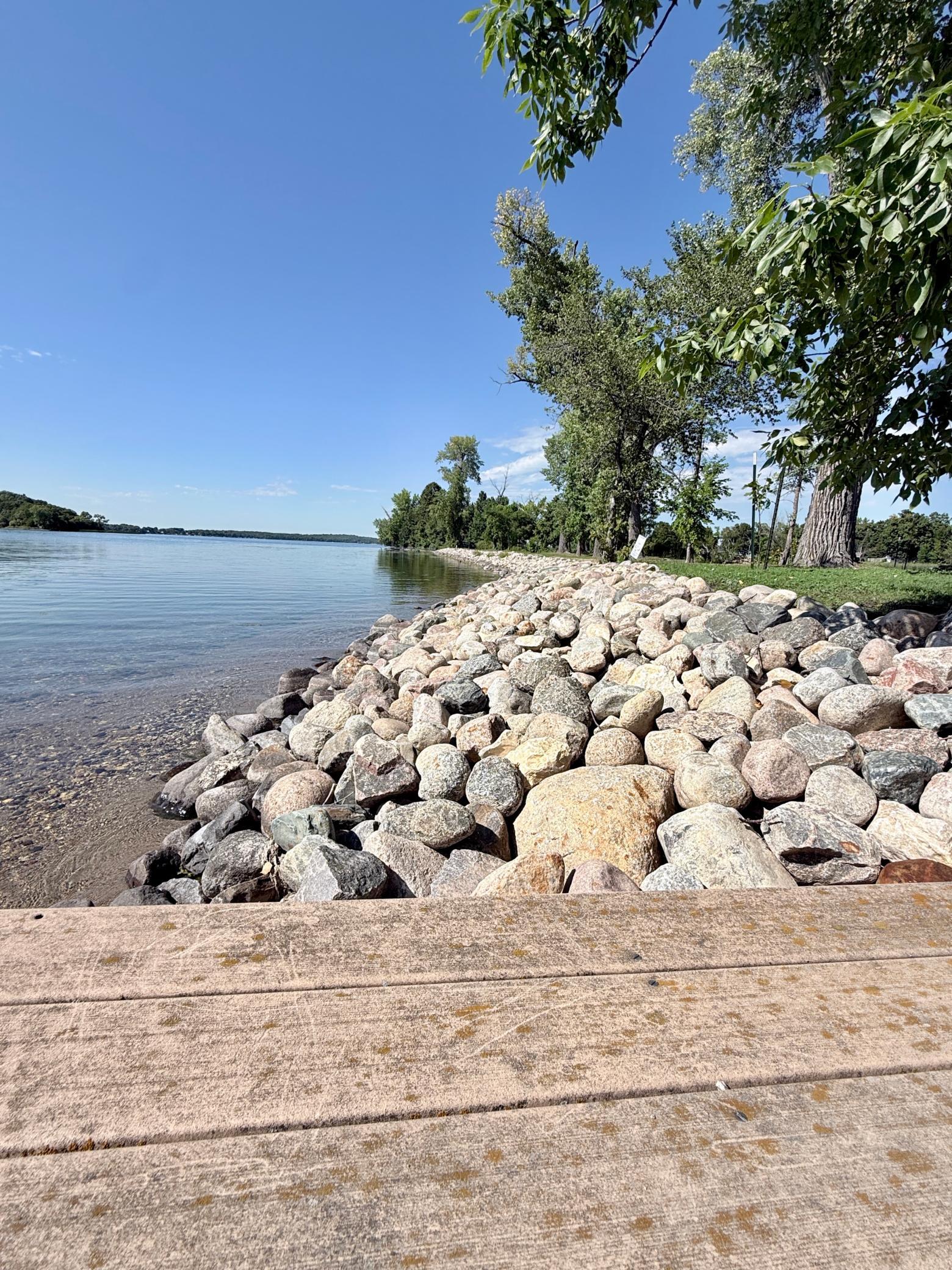 Rock-armored lakeshore beside calm water, shaded by mature trees.