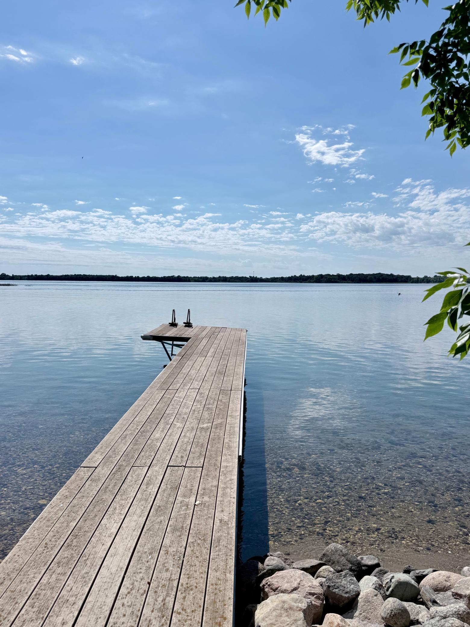 Wooden dock extending into a calm lake, with clear shallow water and rocks along the shore.