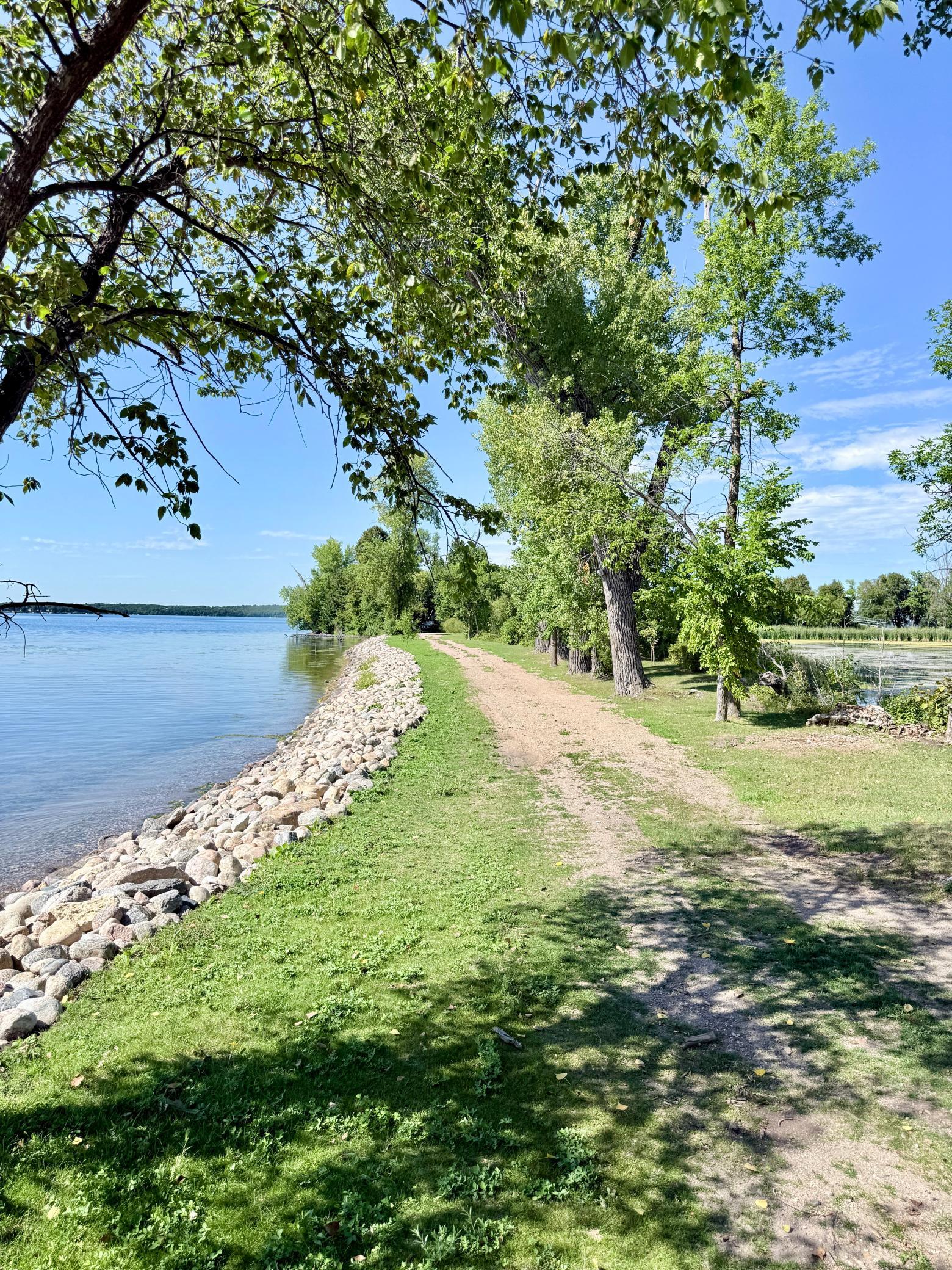 Tree-lined dirt path follows a rock-reinforced lakeshore beside calm water.