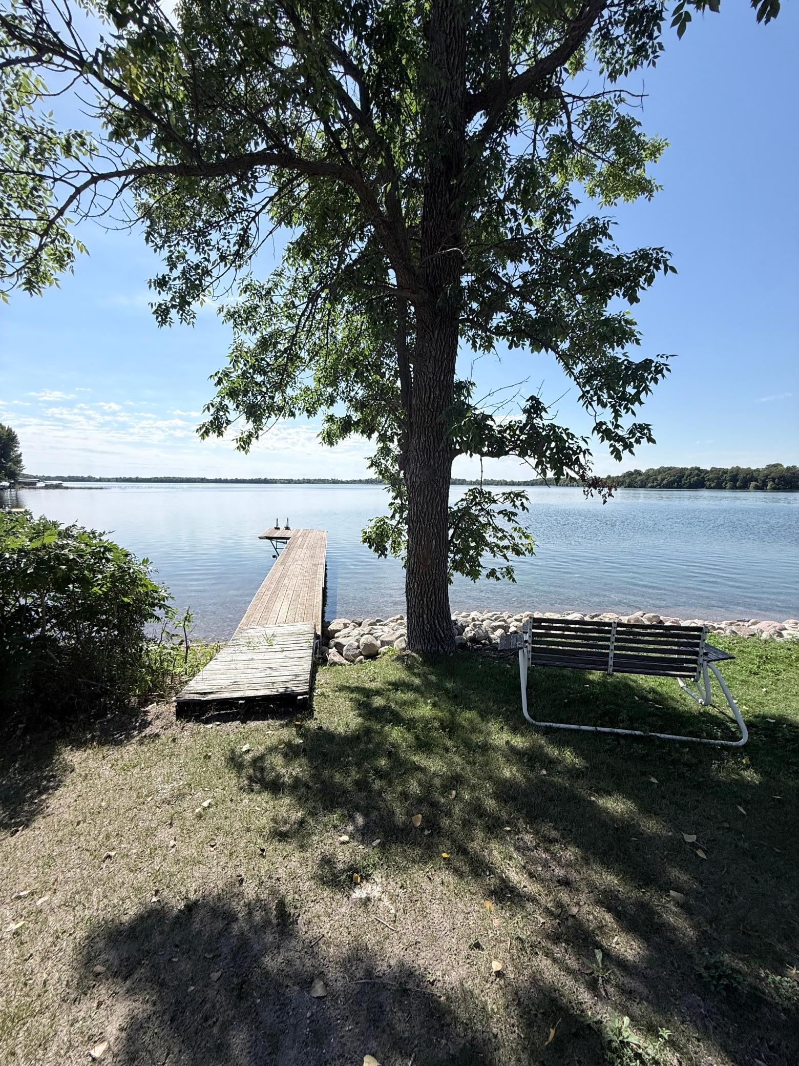 Lakefront yard with a wooden dock, large shade tree, and bench beside the water.