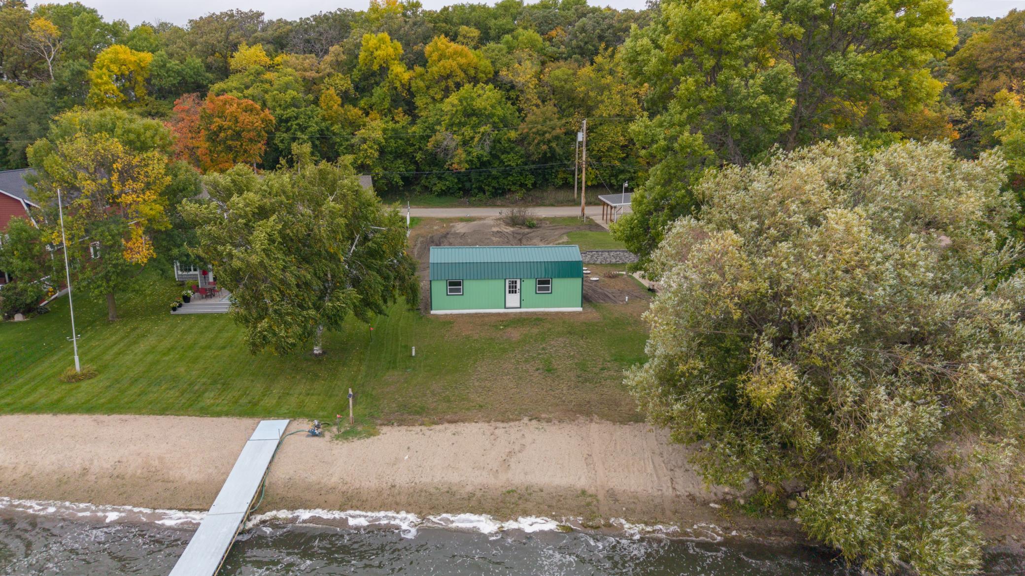 Aerial view of a lakeside property with a sandy beach, dock, and green outbuilding.