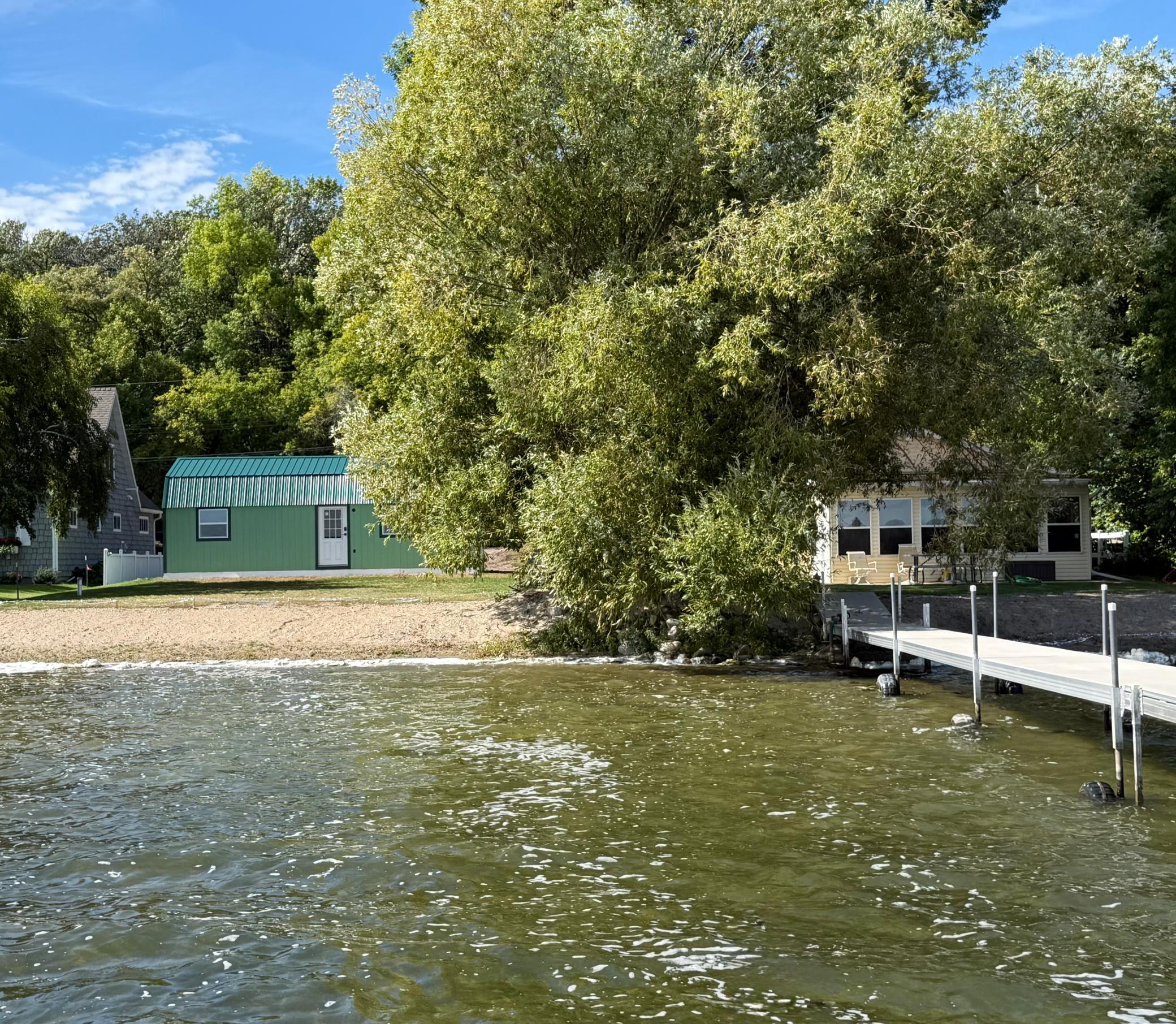 Lakefront home partly shaded by a large tree, with a sandy shore, green outbuilding, and dock.