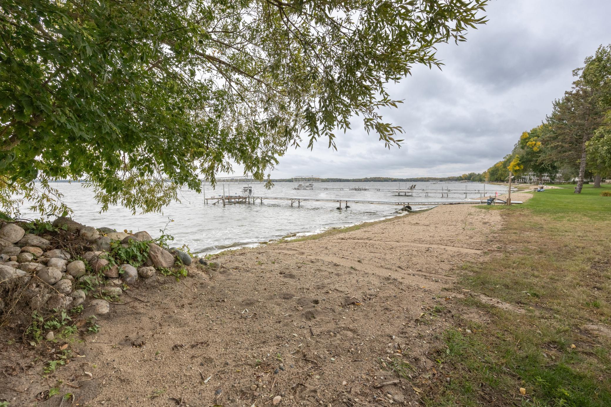 Sandy lakeshore with docks extending into the water beneath leafy trees.