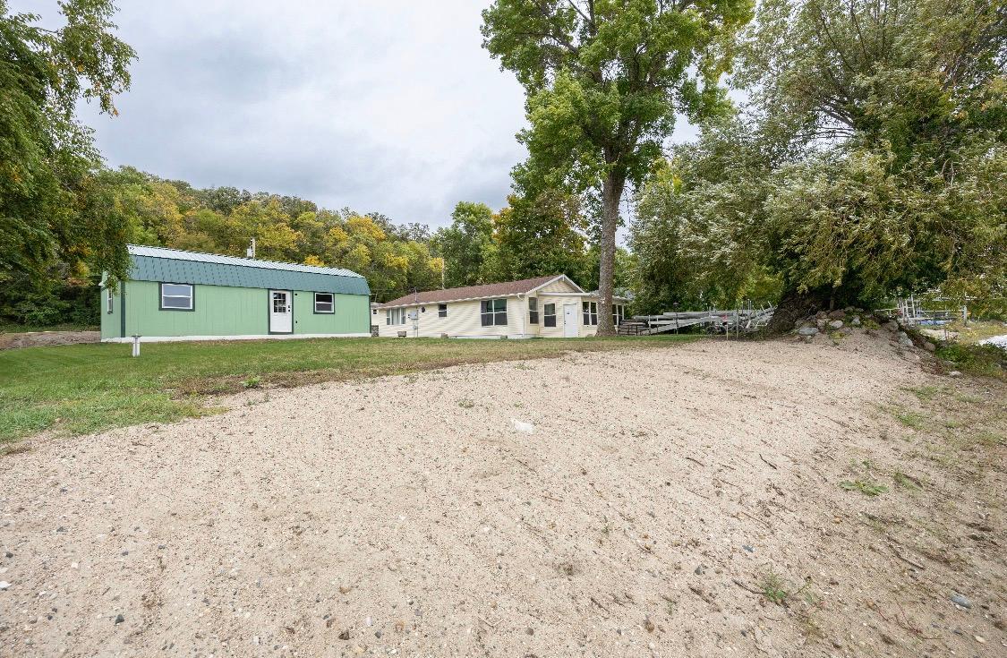 House and green outbuilding beside a broad sandy yard, with mature trees and a dock near the water.