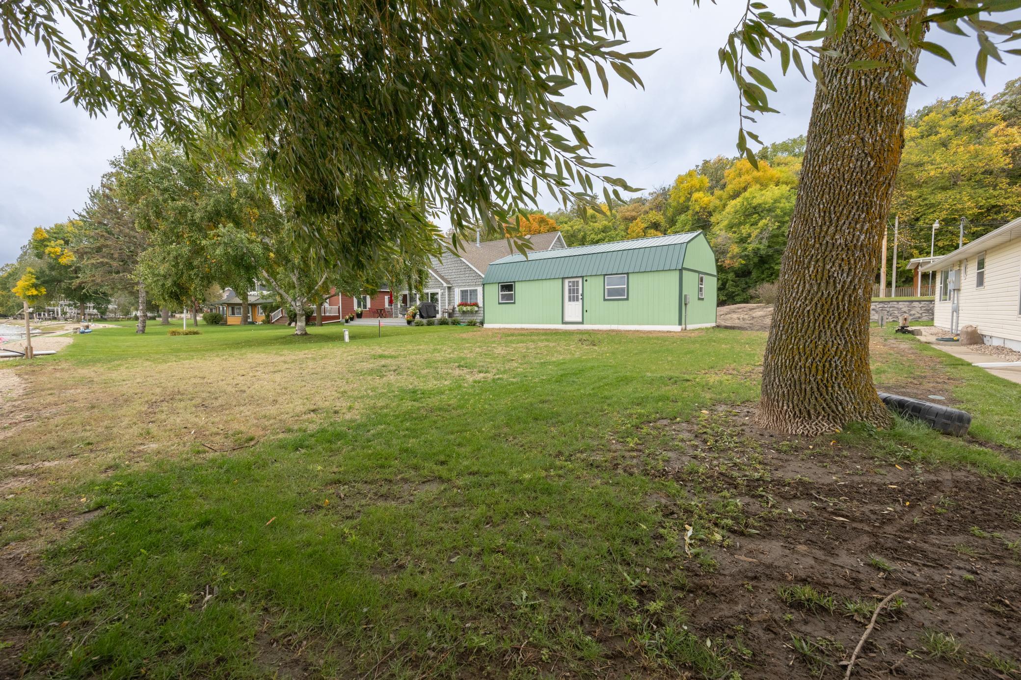 Lakeside property with a green outbuilding, broad lawn, mature trees, and neighboring homes.