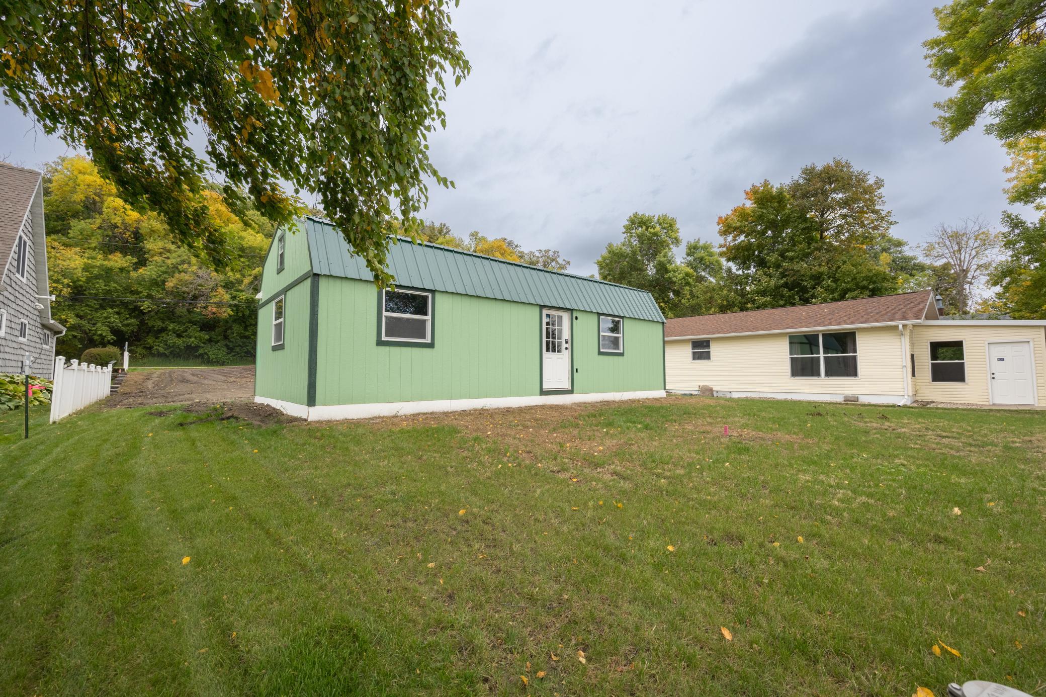 Green metal-sided building beside a beige home in a grassy, tree-lined yard.