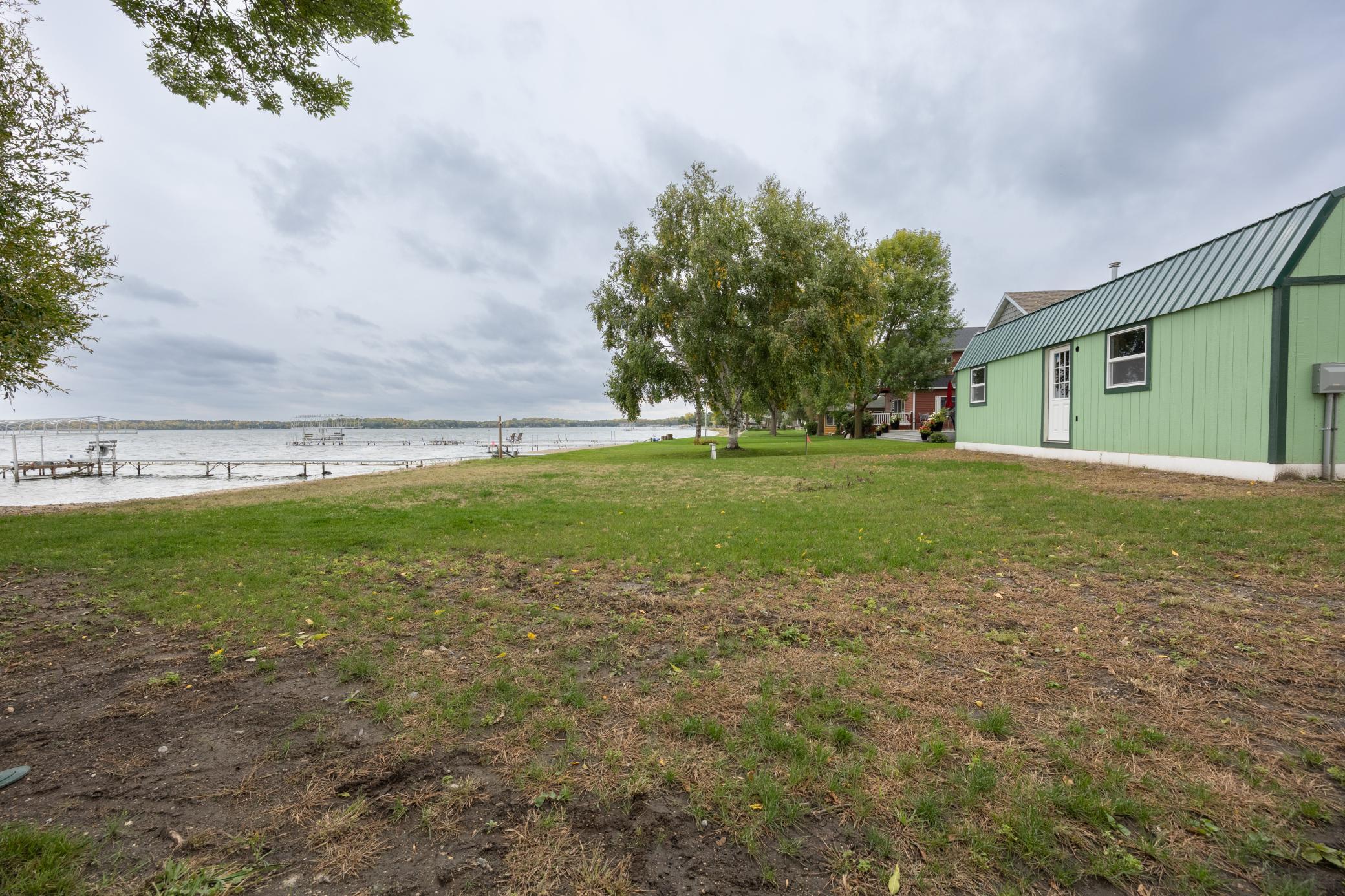 Lakeside yard with a green outbuilding, mature trees, and docks extending into the water.